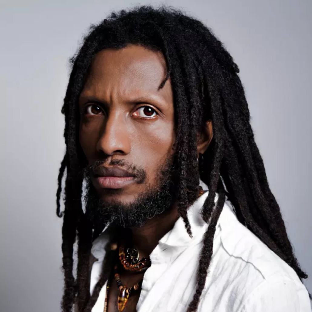 Portrait of Kao Ra Zen. A black man with long black dreads, short beard and mustache, gazing with deep brown eyes into the camera, Zen is wearing a white collared shirt and a beaded colorful necklace.